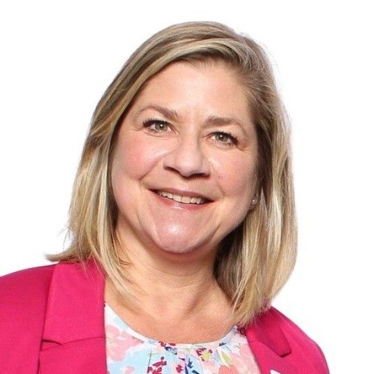 A smiling woman with blonde hair and a pink blazer looks directly at the camera.