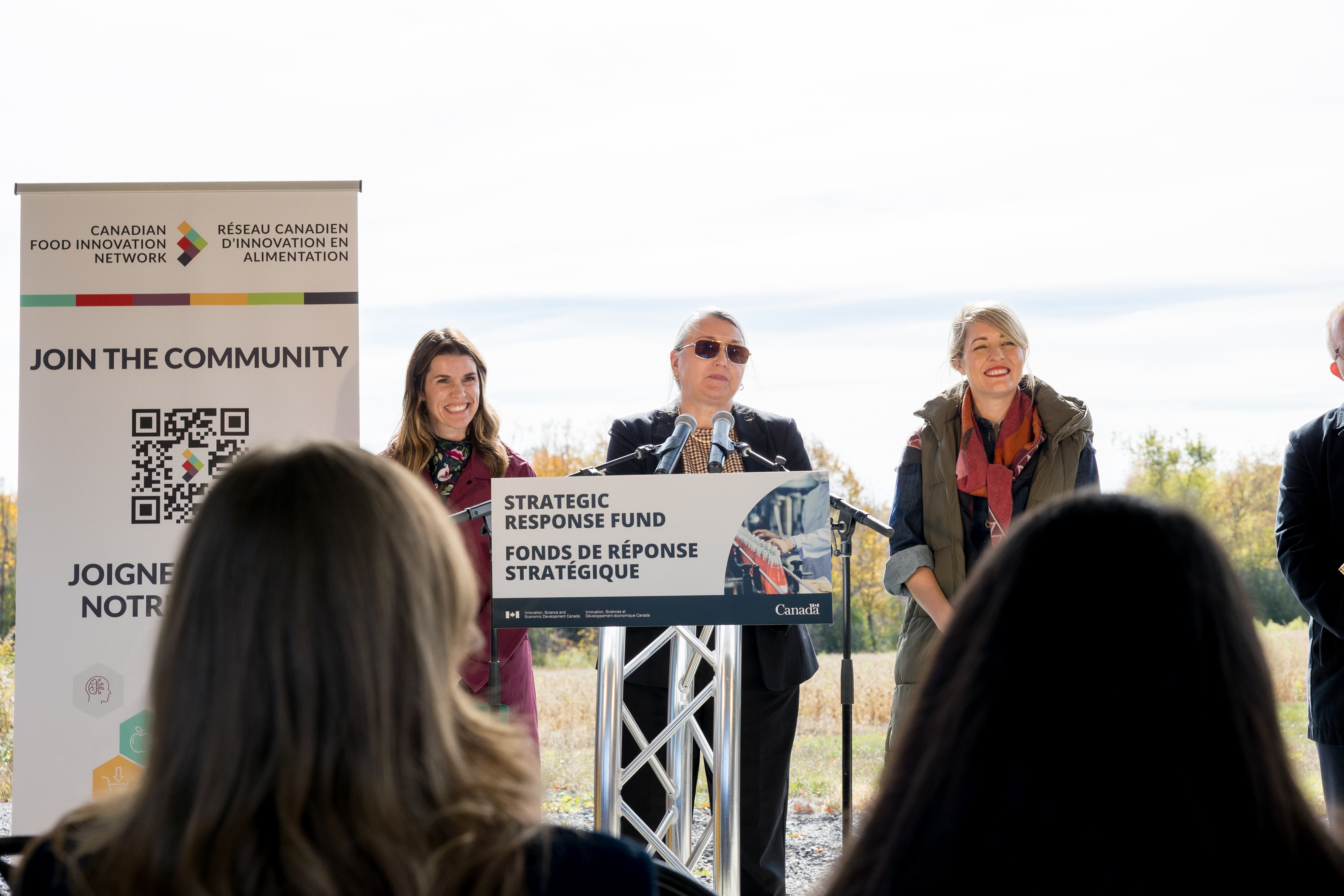 Man speaks at a podium with two women beside him during an outdoor Strategic Response Fund event.