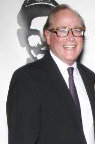 A smiling man in a dark suit and glasses stands against a light background.