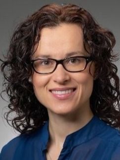 Professional woman with dark curly hair and glasses smiles warmly.