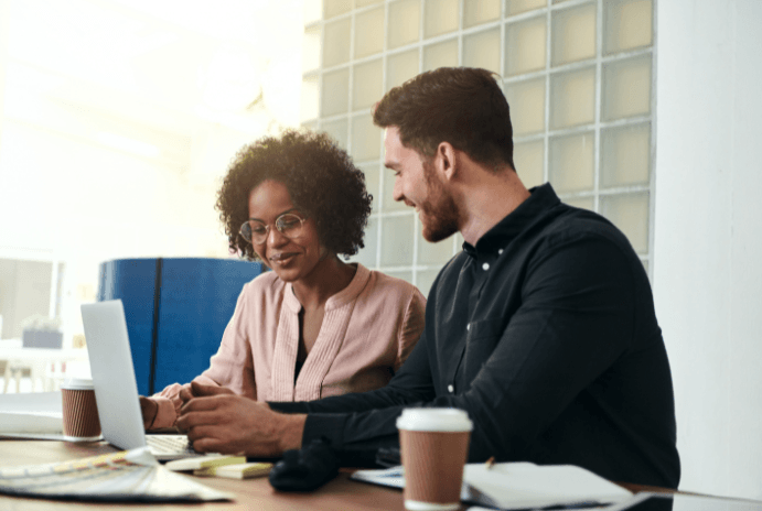 Diverse colleagues collaborate at a bright office desk, looking at a laptop and smiling.