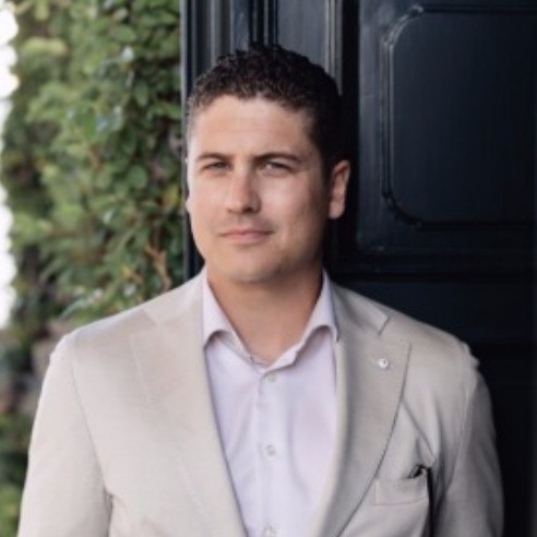 Man in a light suit jacket with a thoughtful expression, foliage in background.