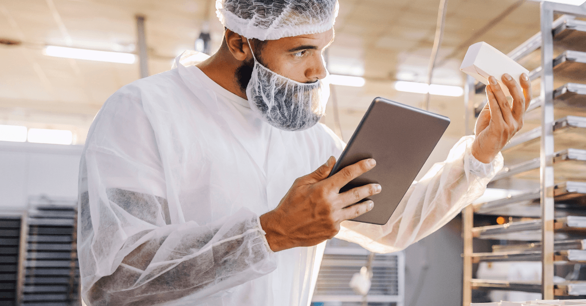 Man in protective suit inspects a product while referencing a tablet in a factory.