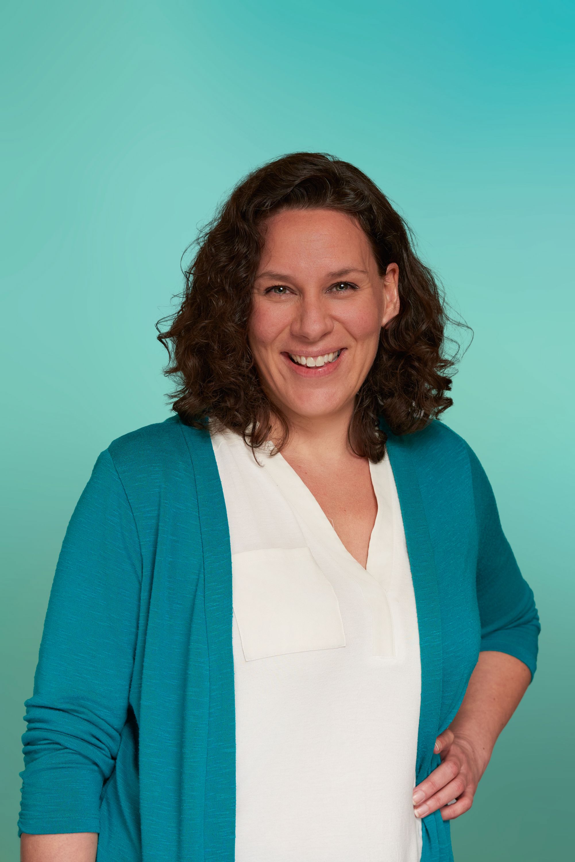 Smiling woman with curly brown hair in white top and teal cardigan against teal background.