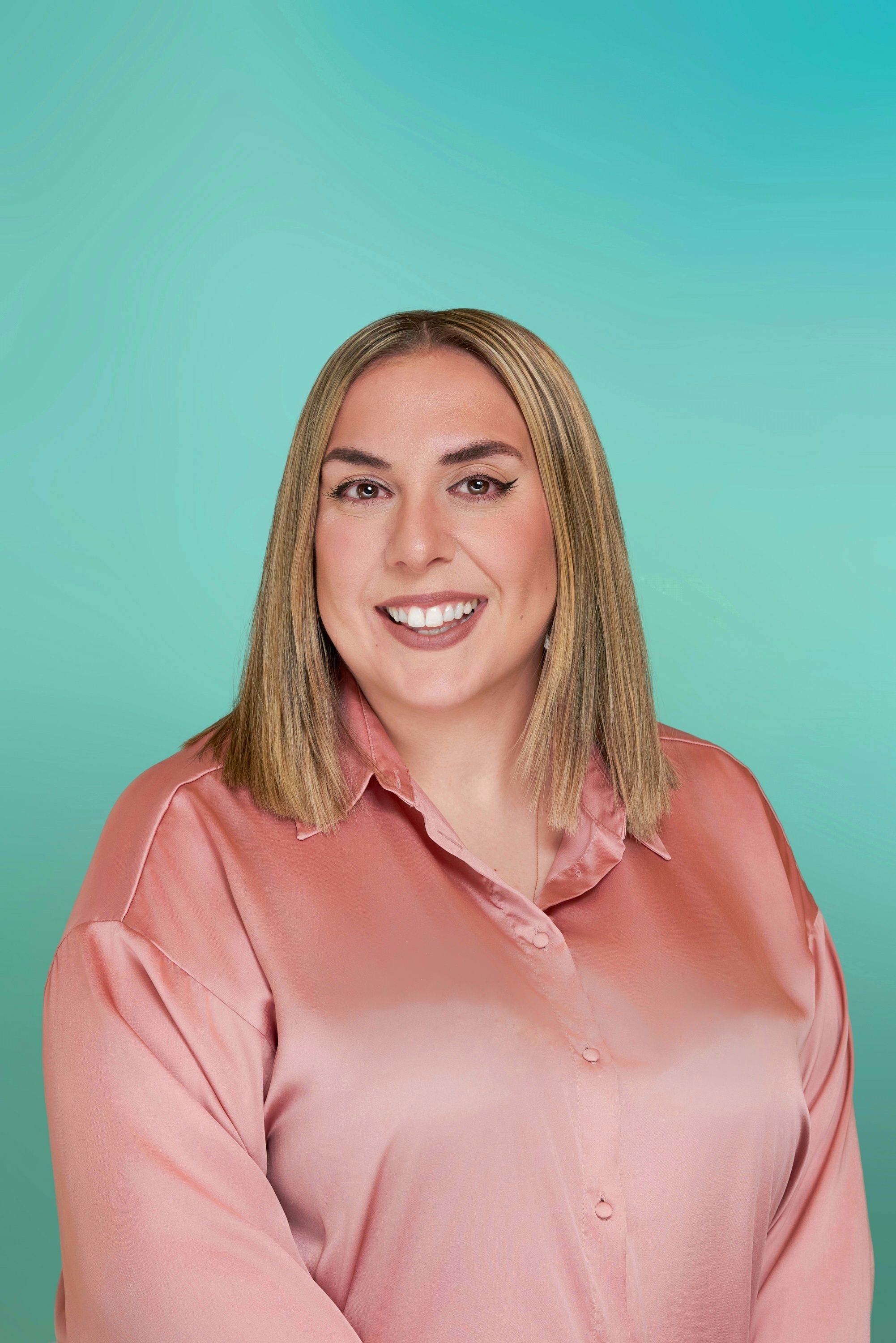 A smiling woman with blonde hair wearing a pink shirt against a teal background.