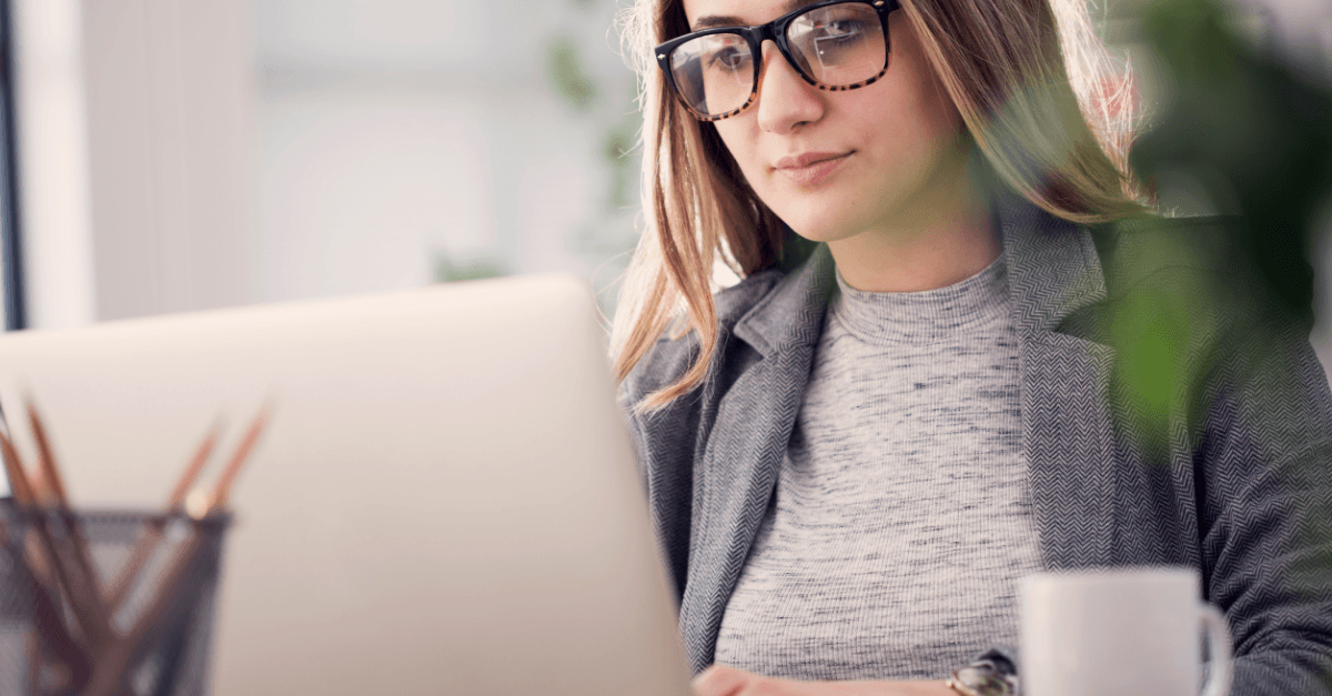 A focused young woman in glasses works diligently on her laptop at a desk.