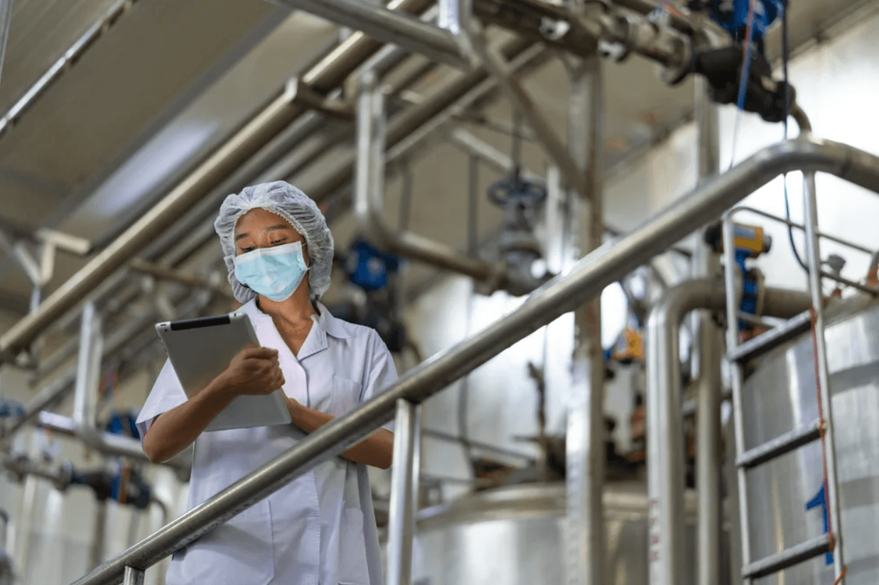 Woman in mask and uniform inspects machinery in a sterile industrial plant.