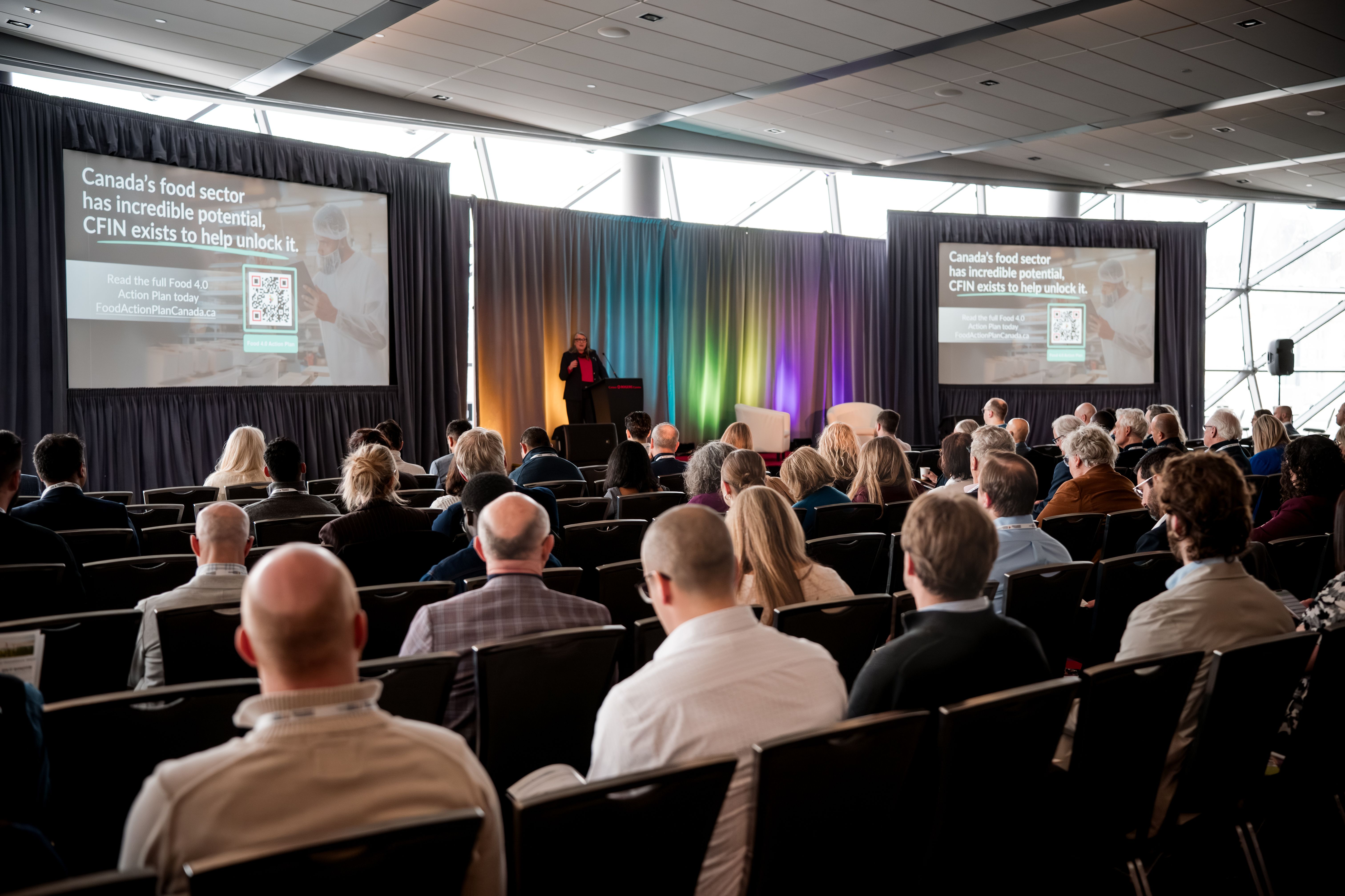 Audience at a conference watching a speaker present on stage with two large screens.