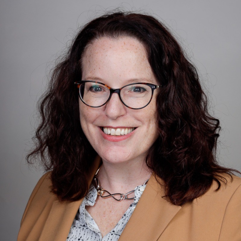 A smiling woman with dark curly hair and glasses wears a tan blazer.