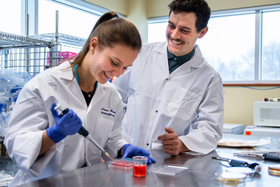 Two smiling scientists in lab coats conduct an experiment with a pipette and petri dish.