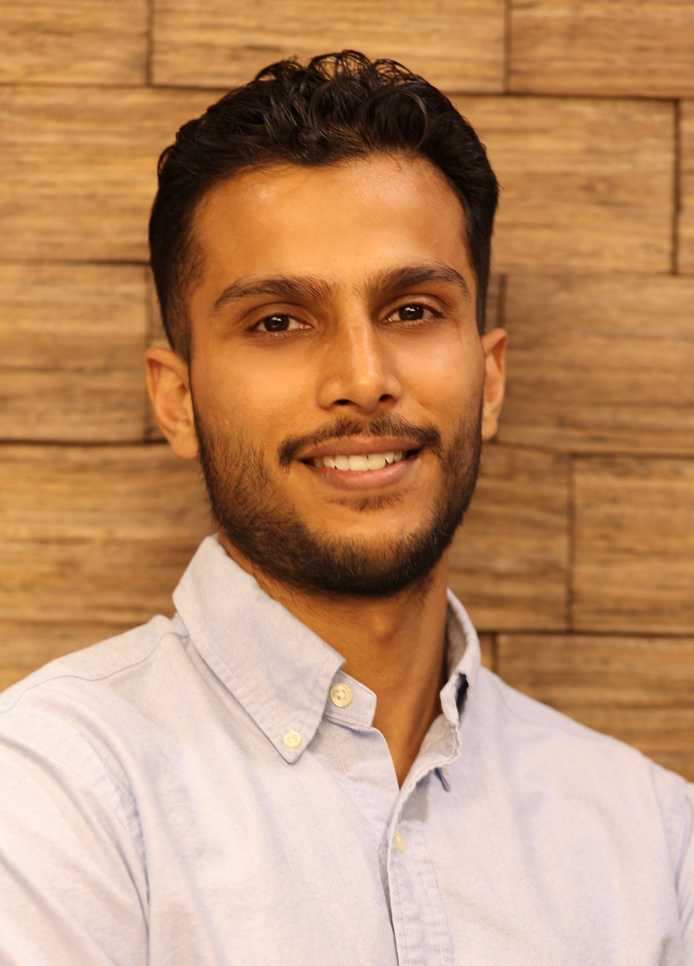 Smiling young man with dark hair and beard in a light shirt against a wooden wall.