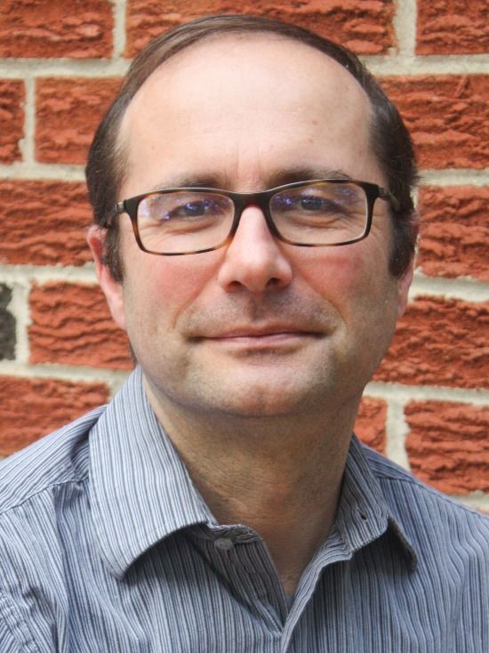 A man with glasses and a striped shirt smiles against a brick wall.