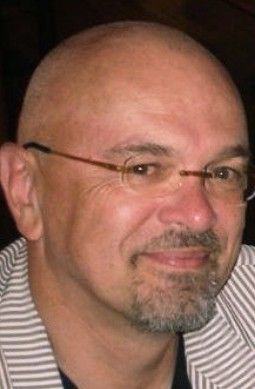 A bald man with glasses, wearing a striped jacket, smiles softly at the camera.