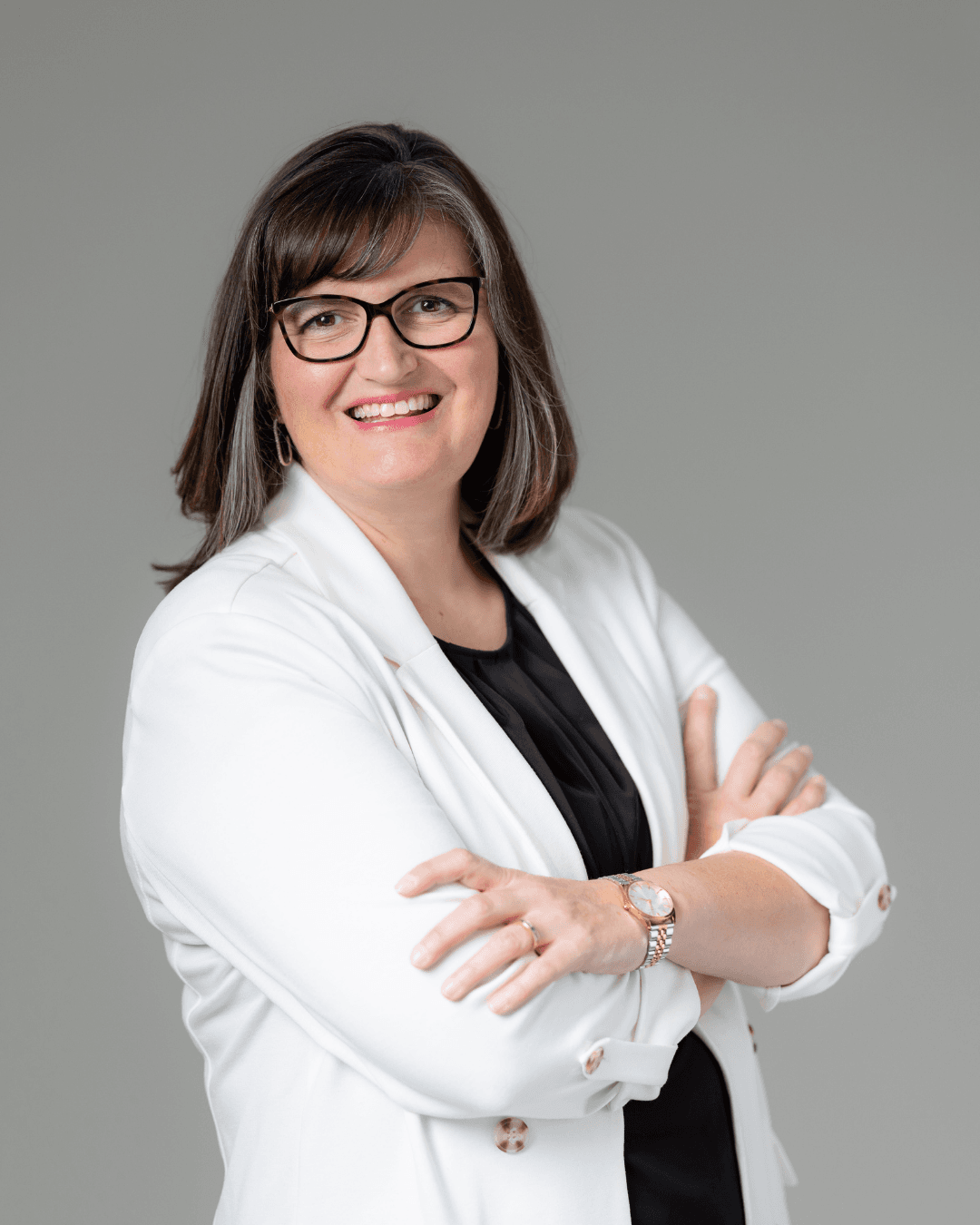 Smiling woman in glasses and white blazer with arms crossed against a gray background.