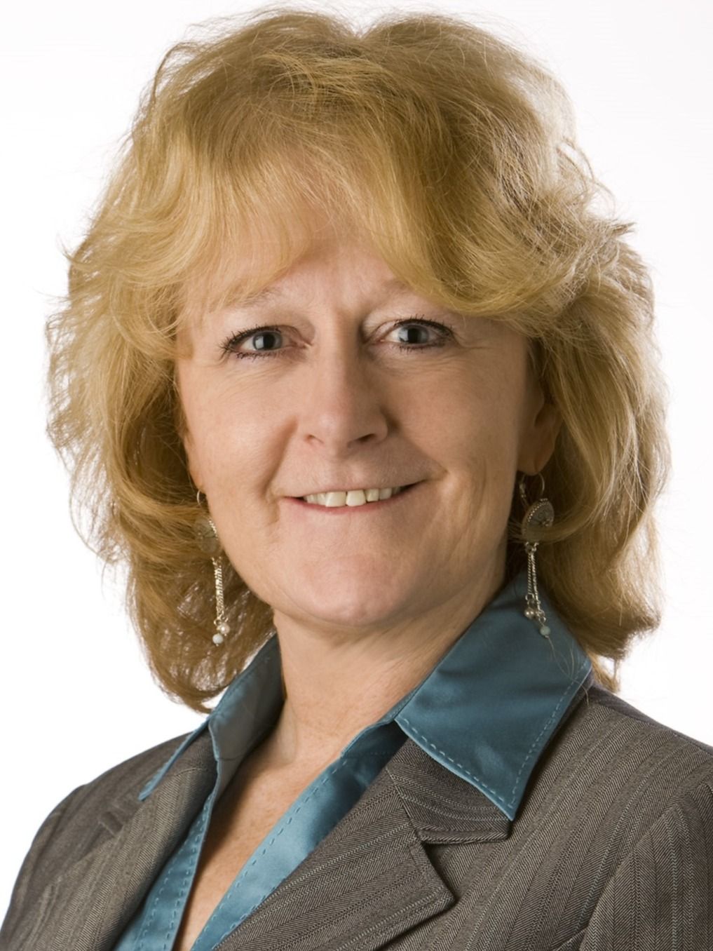Middle-aged woman with blonde curly hair smiling faintly in a grey suit and teal top.