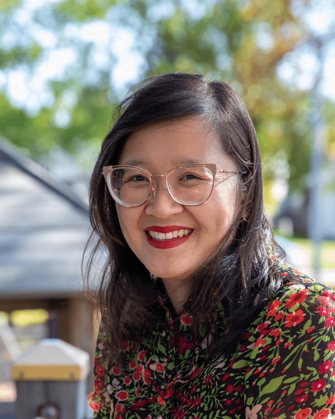 Smiling woman with glasses and dark hair wearing a floral top outdoors.