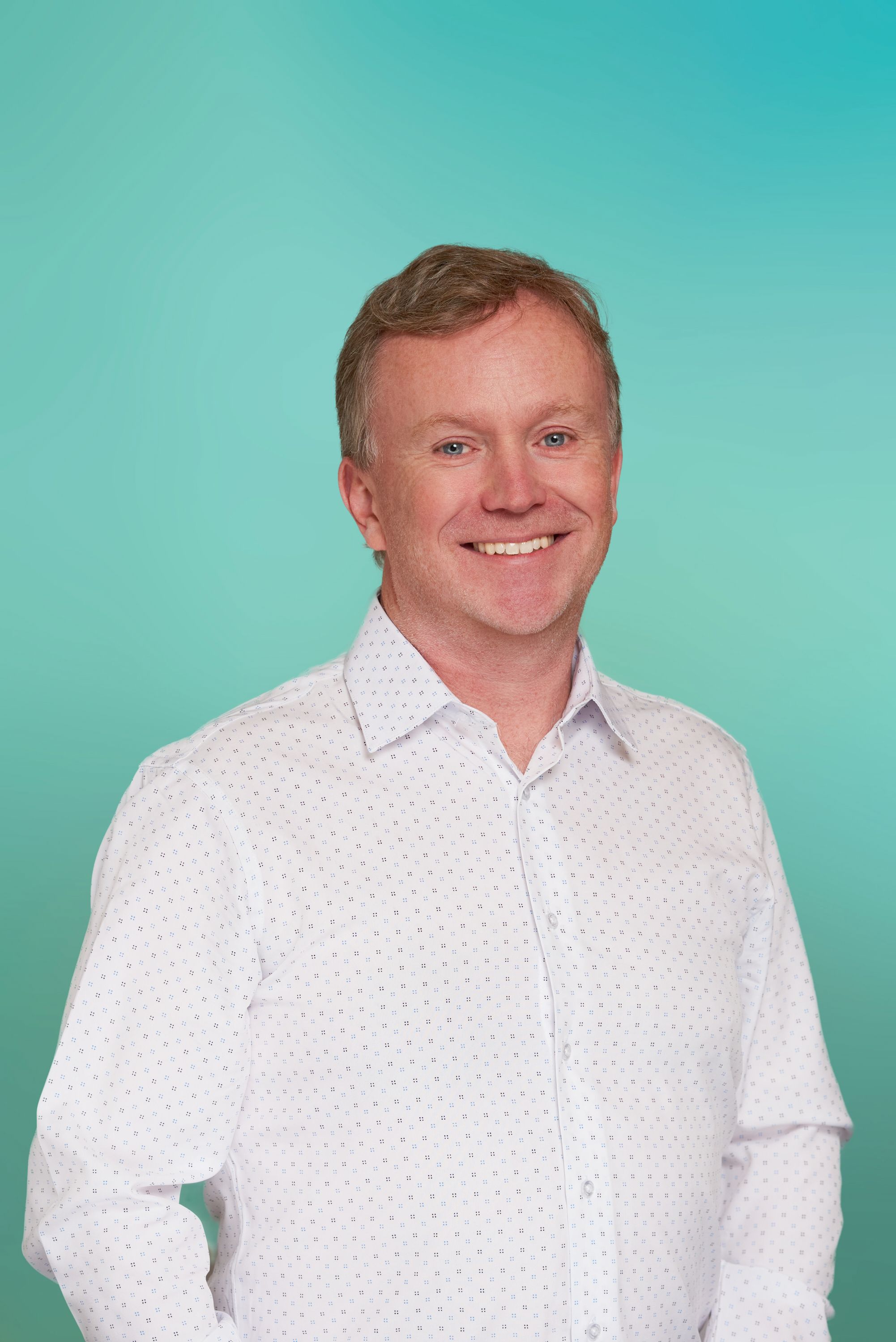 Smiling man with short reddish hair wearing a white shirt against a teal background.