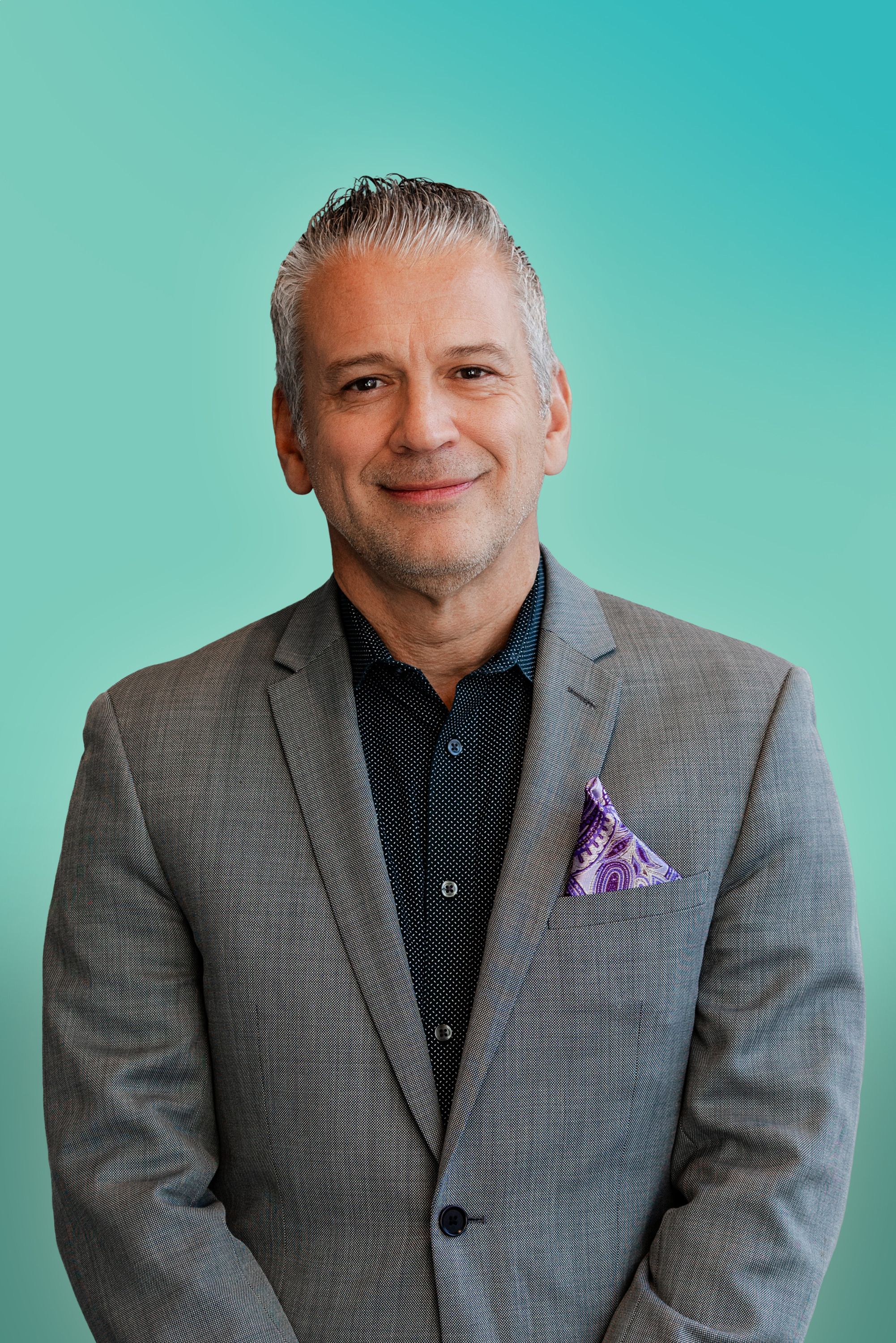 Smiling man in a grey suit and black shirt with a purple pocket square.