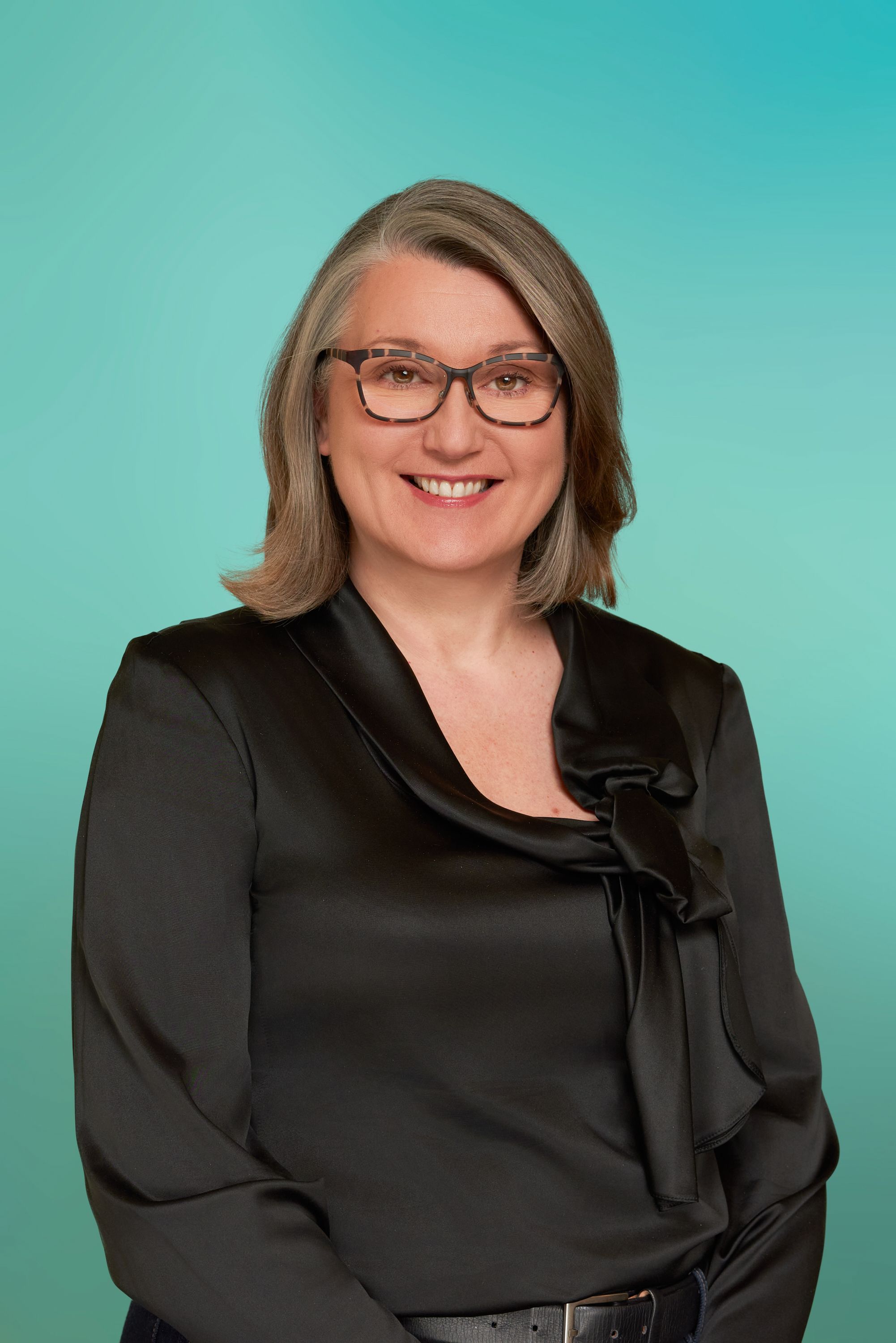 Smiling woman with grey hair and glasses in a black blouse against a teal background.