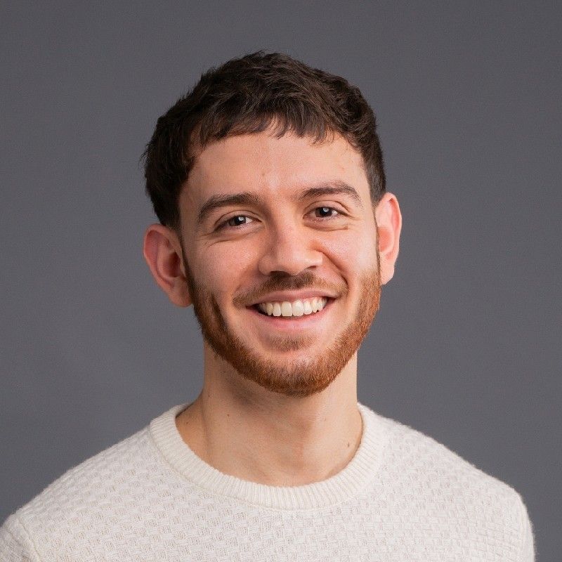 Young man with light brown curly hair and beard smiling against a gray background.