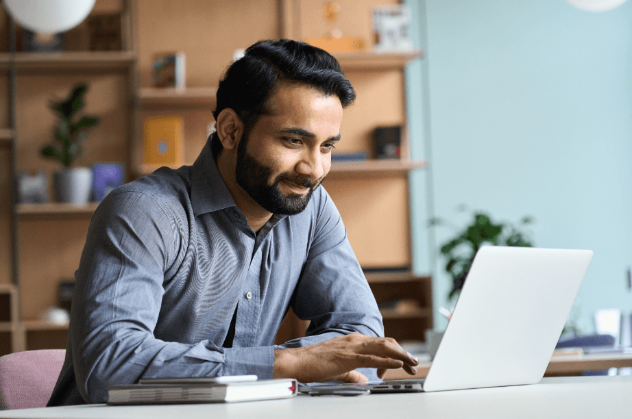 Smiling man in a grey shirt typing on a silver laptop in an office.