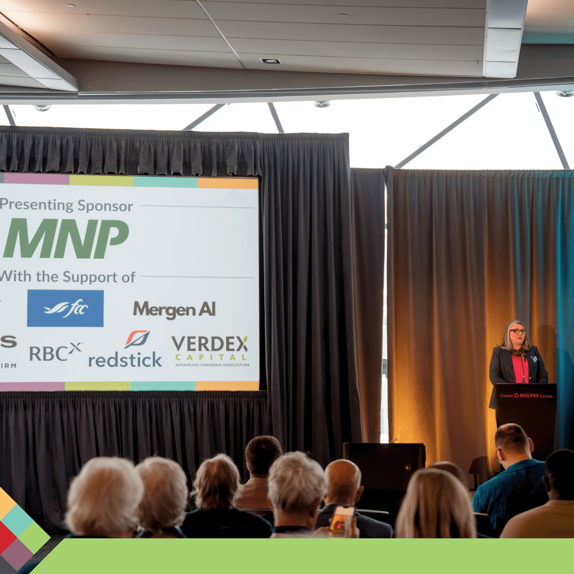 Speaker addresses audience from a podium, screen shows MNP and sponsor logos.
