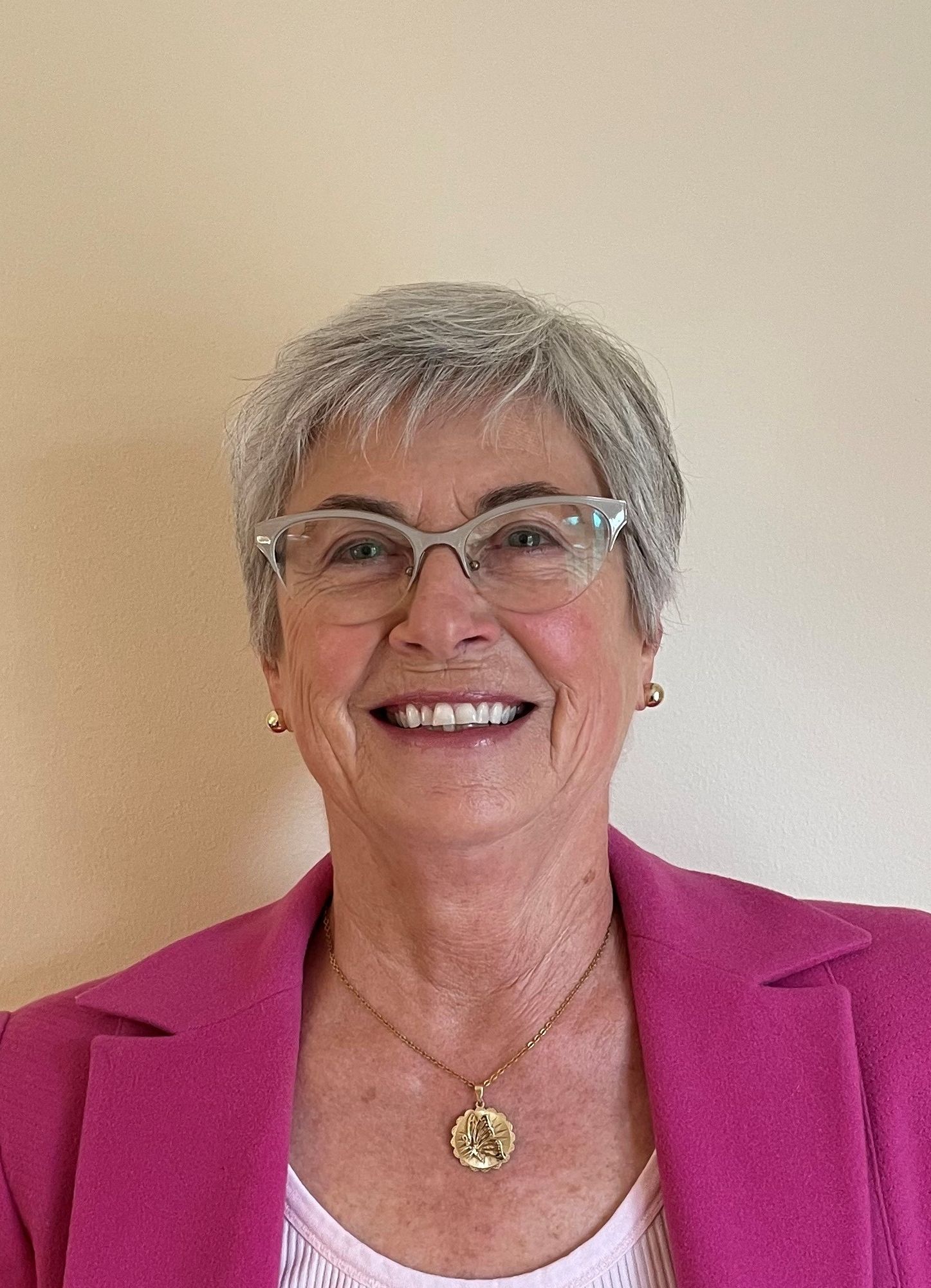 Smiling woman with short grey hair, glasses, and a vibrant pink jacket.