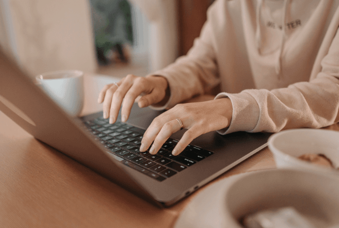 Person wearing a light hoodie typing on a silver laptop with a white mug and snack.