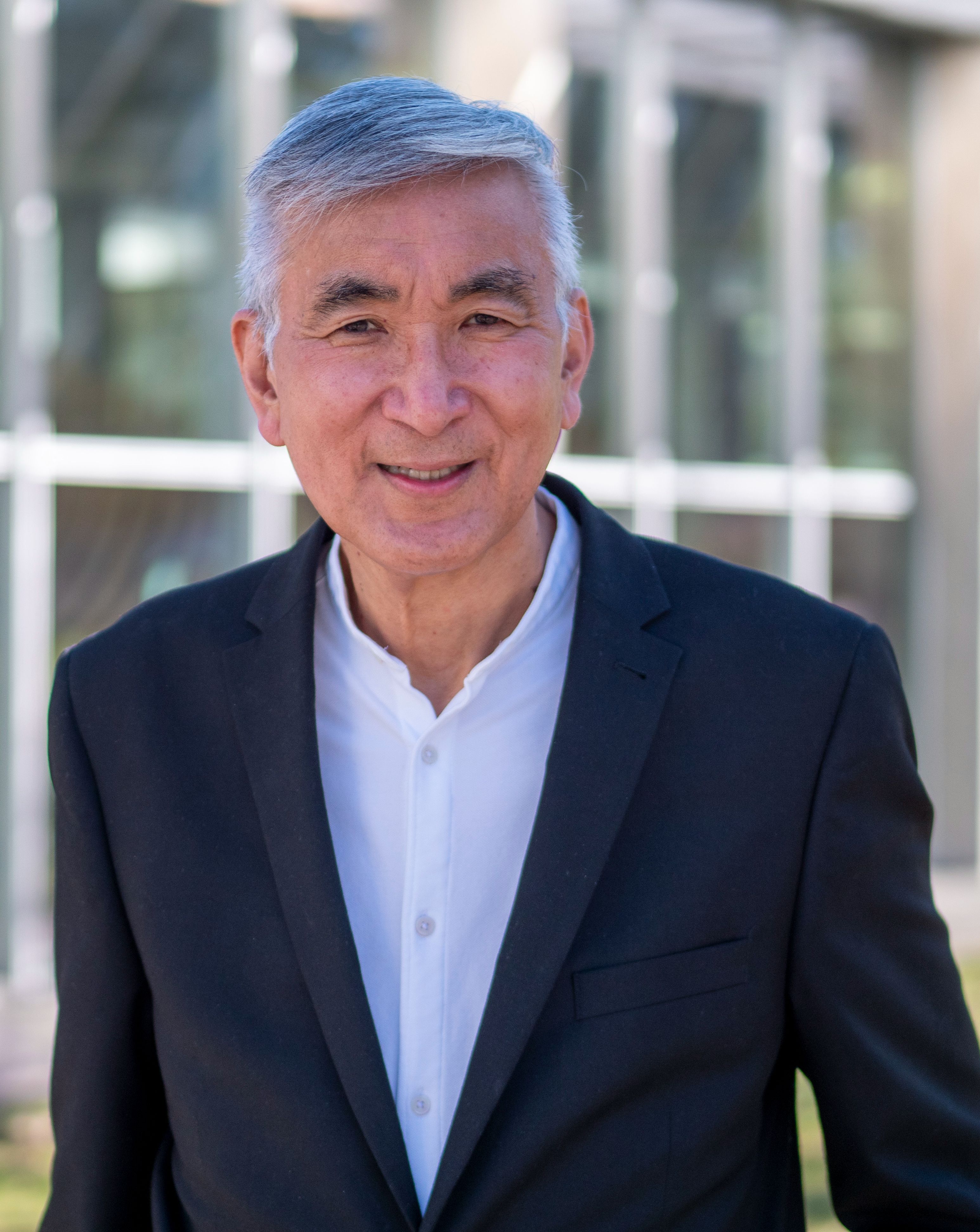 Smiling older Asian man in a dark suit jacket and white shirt outdoors.