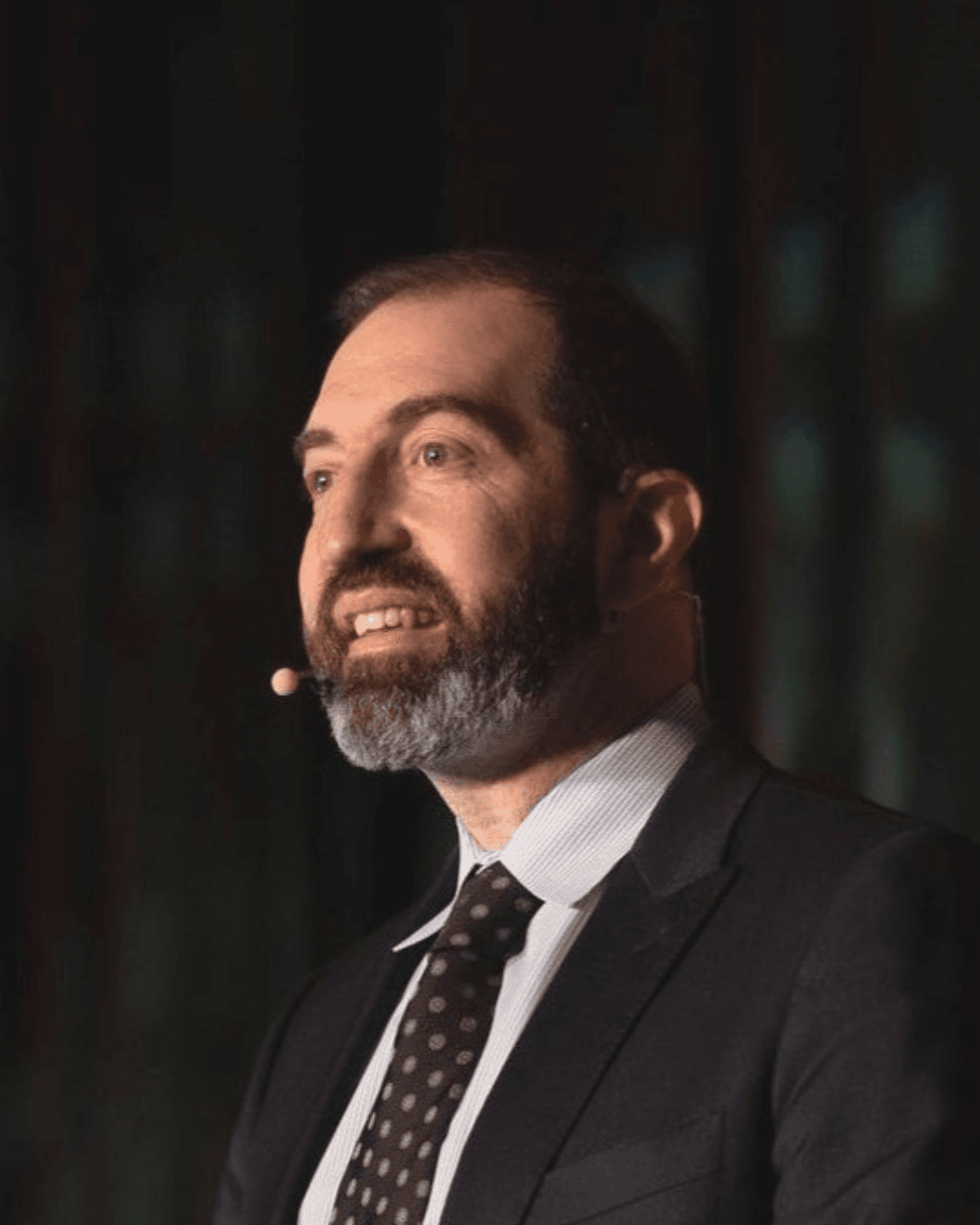 Man with beard speaking into a lapel microphone during a professional presentation.