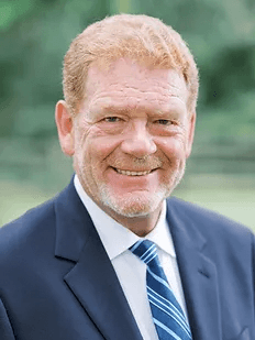 A smiling man with red hair and beard in a dark suit and blue striped tie.