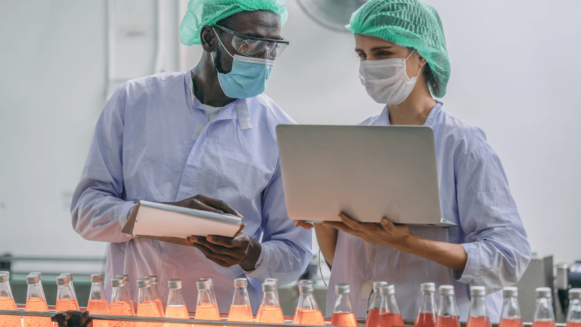 Two workers in protective gear inspect a beverage production line using a laptop and clipboard.