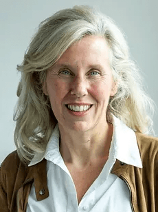 Smiling woman with wavy grey hair, blue eyes, wearing a white shirt and brown jacket.