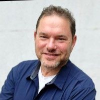 Man with short dark hair and a beard wearing a blue shirt, smiling.