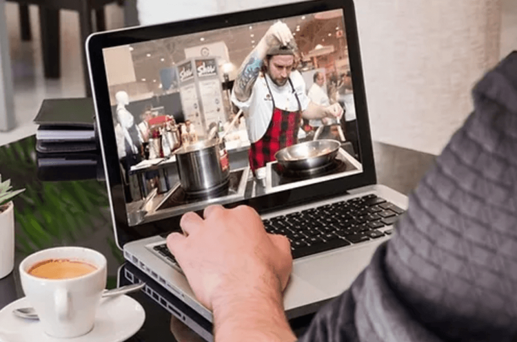 Person watching a chef cook on a laptop while drinking coffee.