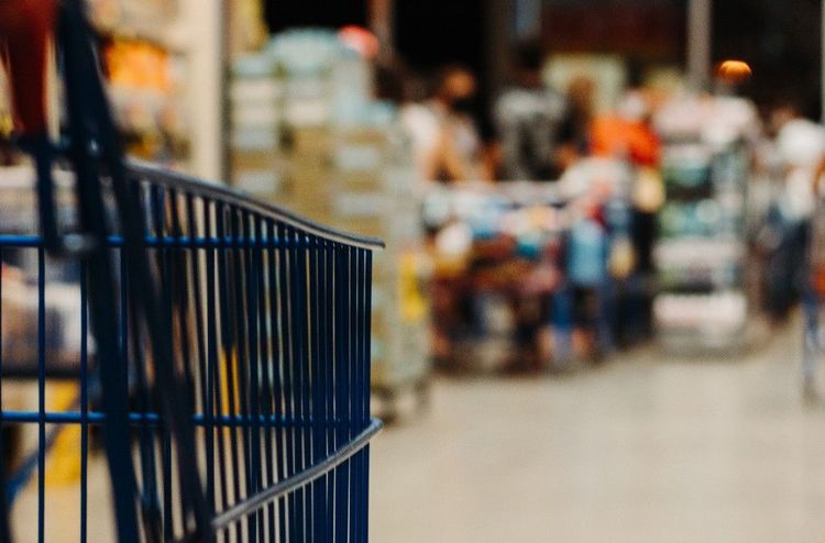 Close-up of a blue shopping cart in a blurred supermarket aisle with other shoppers.
