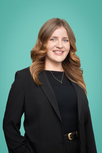 A smiling woman with long brown hair wears a black blazer against a teal background.