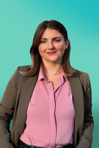 Smiling woman with brown hair wearing a pink shirt and dark blazer against a teal background.