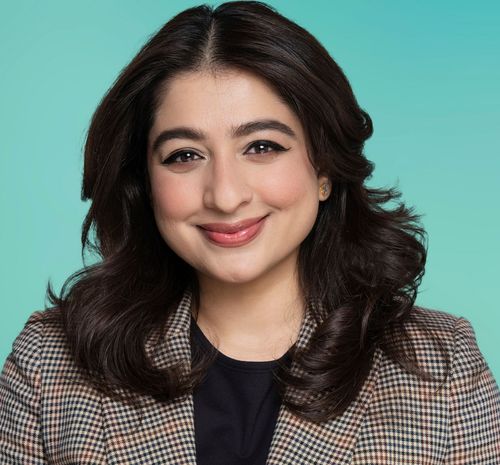 A smiling woman with dark hair wears a plaid blazer against a teal background.