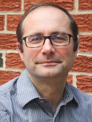 A man with glasses and a striped shirt smiles against a brick wall.