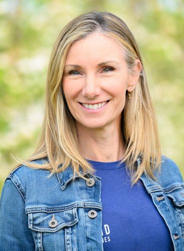 A smiling woman with blonde hair and a denim jacket against a blurred green background.