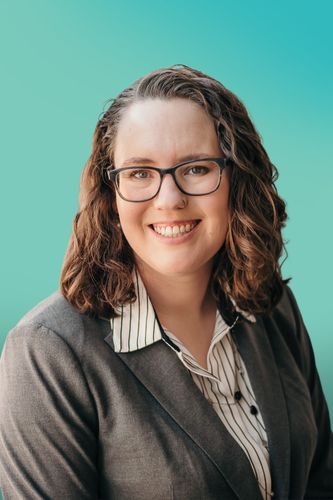 Smiling woman with dark curly hair and glasses wearing a professional blazer.