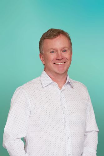 Smiling man with short red hair wearing a white button-down shirt against a teal background.