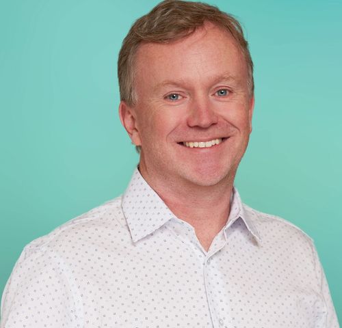 Smiling man with red hair in a patterned shirt against a teal background.