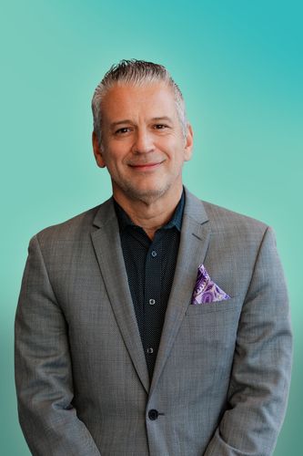 Smiling man in a grey suit and black shirt with a purple pocket square.