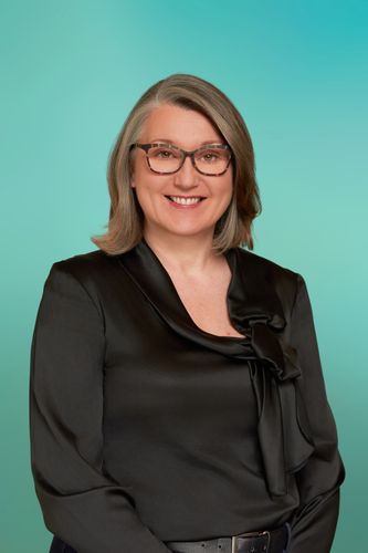 Smiling woman with glasses and gray hair, wearing a black satin blouse.