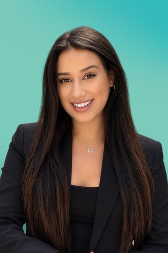 Smiling young woman with long dark hair wearing a black blazer against a teal background.